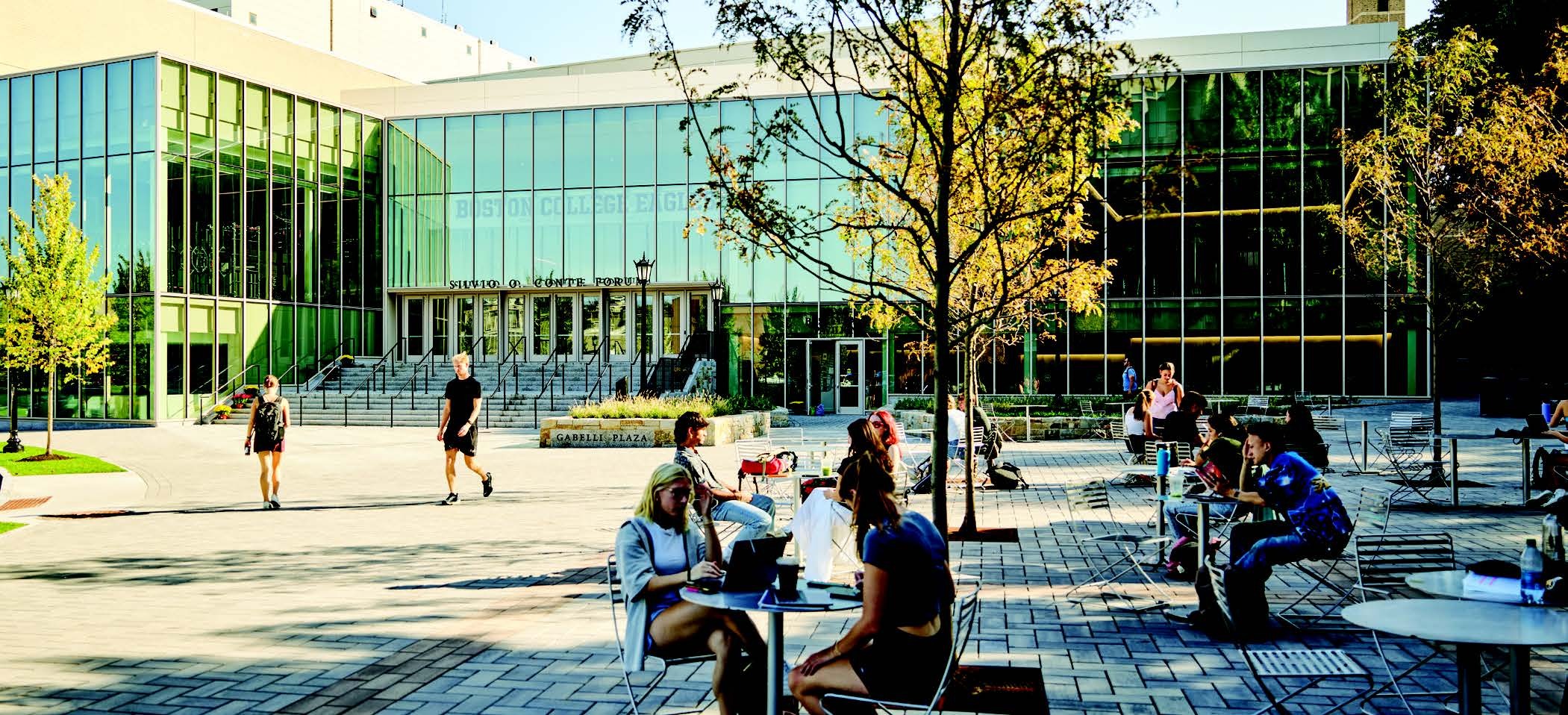 A modern glass-fronted building with "Harold B. Lee Library" written above the entrance. In front, people are sitting at tables, chatting, reading, and using laptops. The area is adorned with trees and paved with bricks, creating a lively and inviting atmosphere.